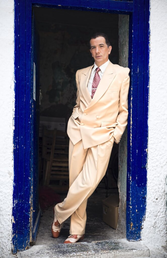 Ολόσωμη φωτογραφία του Ariel Perez να στέκεται στο μπλε κάδρο μιας πόρτας. Φοράει ένα μπεζ κοστούμι και παπούτσια tango.