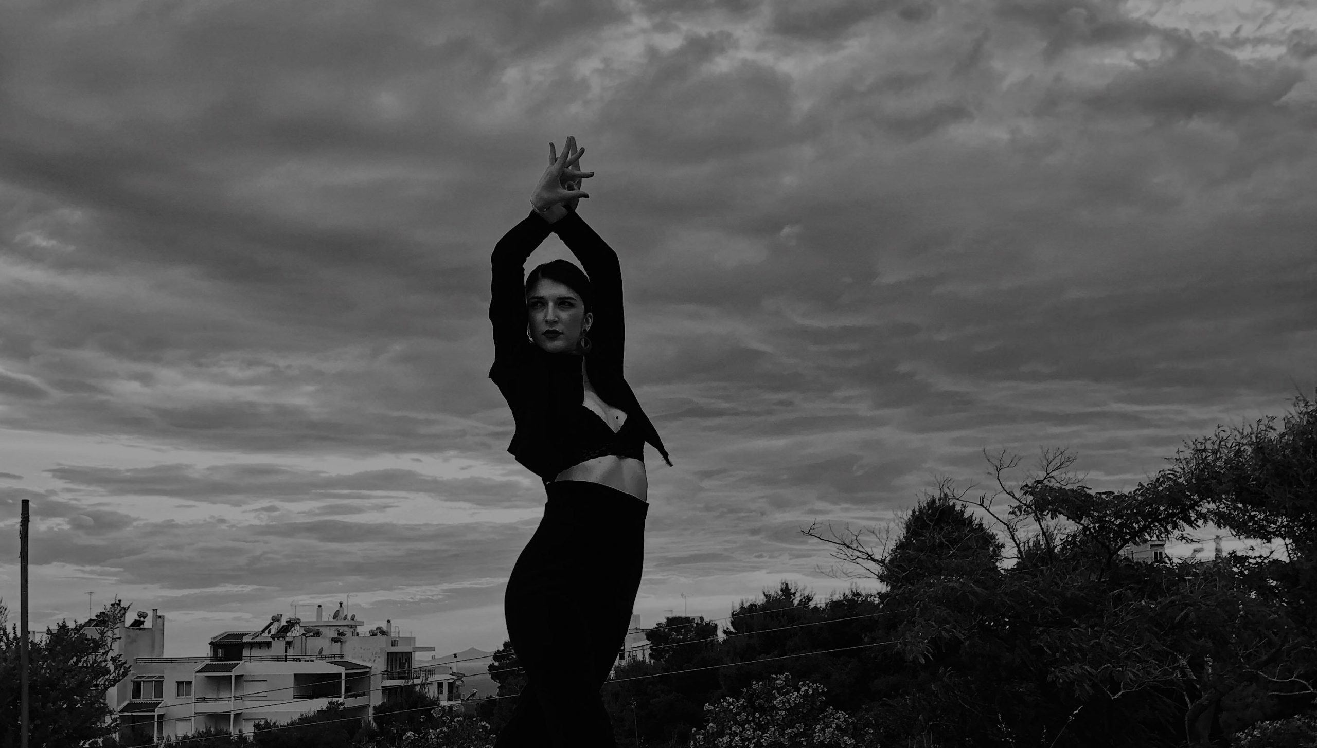 Φωτογραφία της Βάσια Κατσιγιάννη που κάνει μια πόζα flamenco με τα χέρια της προς τα πάνω. Φοράει μαύρα ρούχα και πίσω της φαίνεται ο συννεφιασμένος ουρανός.