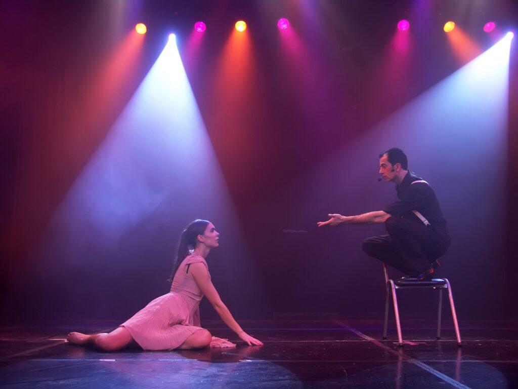 Ο Ariel Perez και η Margarita Plessa κοιτάζονται σε μια σκηνή θεάτρου, φωτισμένη από ροζ φως. Ο Ariel πατάει σε μια καρέκλα και η Margarita είναι στο πάτωμα.