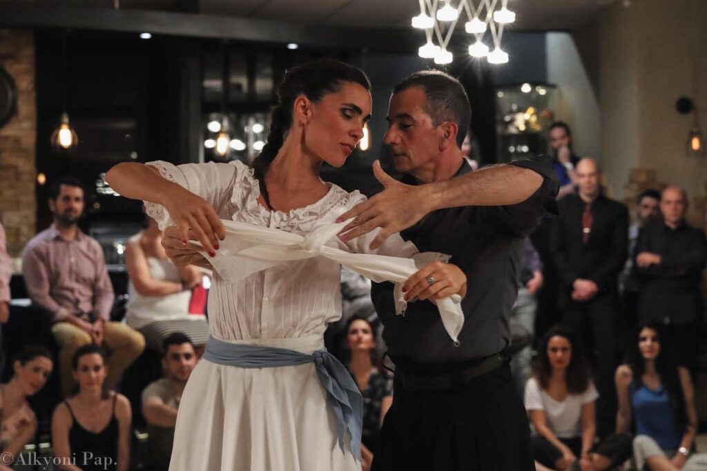 Ο Ariel Perez και η Margarita Plessa χορεύουν zamba κρατώντας μαντίλια στα χέρια τους. Πίσω τους υπάρχει κόσμος που τους κοιτάζει.