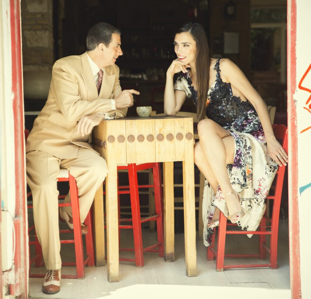 Φωτογραφία των Ariel Perez και Margarita Plessa, ντυμένων με ρούχα tango να κάθονται σε ένα ξύλινο τραπεζάκι σε ένα μαγαζί και να χαμογελάνε