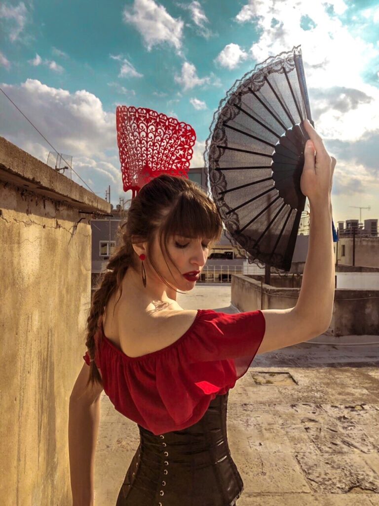 Φωτογραφία προφίλ της Βάσια Κατσιγιάννη που κάνει μια κίνηση flamenco με μια βεντάλια. Φοράει ένα μαύρο-κόκκινο φόρεμα flamenco και στέκεται σε μια ταράτσα.