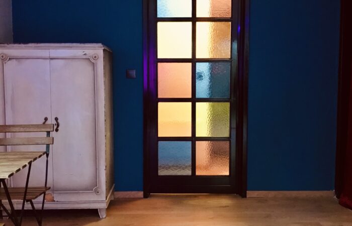 φωτογραφία από το εσωτερικό του Tango Atelier Home. Φαίνεται ένας μπλε τοίχος με μια γυάλινη πόρτα με πολύχρωμα βιτρό και ένα τραπέζι στο πλάι.